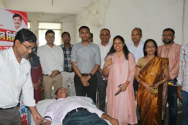 अस्पताल के कमरे में बिस्तर पर लेटा एक व्यक्ति रक्तदान कर रहा है और आसपास अधिकारी व अन्य लोग खड़े हैं