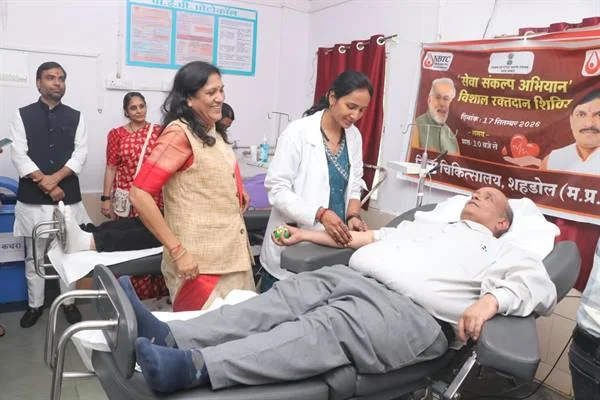 अस्पताल के रक्तदान कक्ष में कुर्सी पर लेटा एक व्यक्ति रक्तदान करते हुए, पास में स्वास्थ्यकर्मी और अन्य लोग खड़े हैं