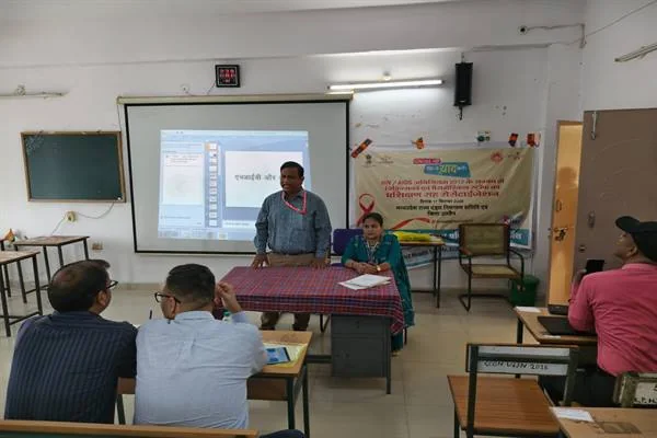 कक्षा में प्रोजेक्टर स्क्रीन के सामने खड़े प्रशिक्षक और मेज पर बैठी महिला, सामने बैठे प्रतिभागी