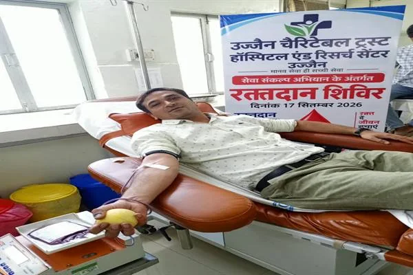 रक्तदान कुर्सी पर लेटा एक व्यक्ति हाथ में पीली गेंद पकड़े हुए रक्तदान कर रहा है और पीछे शिविर का पोस्टर लगा है