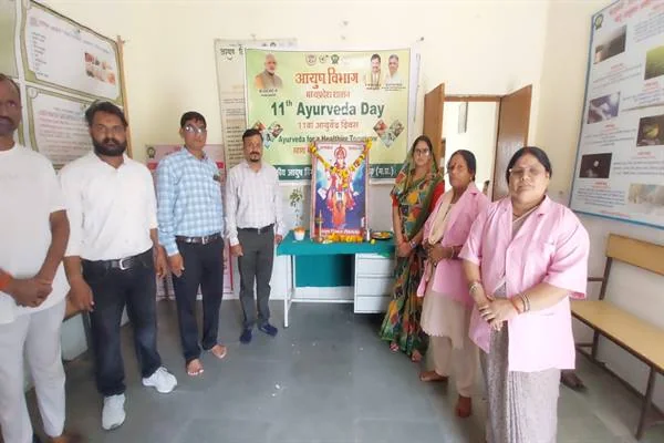 आयुष विंग के कमरे में भगवान धन्वंतरि के चित्र और आयुर्वेद दिवस के बैनर के पास खड़े अधिकारी, चिकित्सक और कर्मचारी