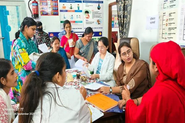 अस्पताल के एक कमरे में मेज के चारों ओर बैठी महिला चिकित्सक और स्वास्थ्यकर्मी कागज़ों पर जांच का ब्योरा दर्ज कर रही हैं, पास में कई महिलाएं अपनी बारी का इंतजार करते हुए खड़ी हैं। पीछे दीवार पर स्वास्थ्य संबंधी पोस्टर और 'डॉक्टर परामर्श' लिखा कागज लगा है।
