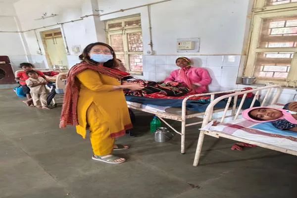 अस्पताल के वार्ड में मास्क पहने एक महिला बिस्तर पर बैठी महिला को सामग्री देती हुईं, आसपास बिस्तरों पर महिलाएं और छोटे बच्चे।