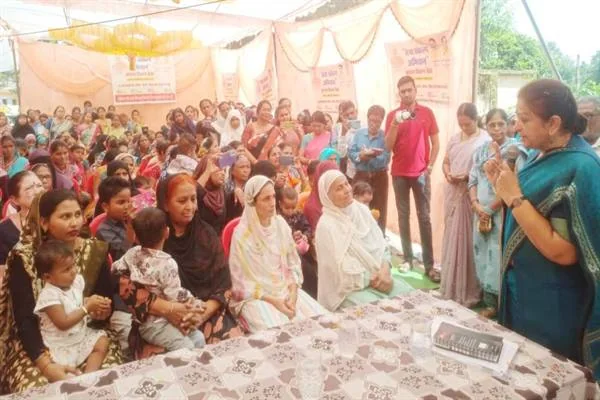 पंडाल में माइक थामे खड़ी एक महिला संबोधित कर रही हैं, सामने जमीन पर बैठी महिलाएं और गोद में बच्चे लिए माताएं सुन रही हैं।