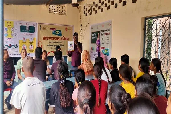 एक कमरे में बेंचों पर बैठे छात्र-छात्राएं और ग्रामीण, सामने खड़ा एक व्यक्ति माइक लेकर बोल रहा है; पीछे दीवार पर 'सघन जागरूकता अभियान' और एचआईवी जांच से जुड़े रंगीन बैनर व पोस्टर लगे हैं।